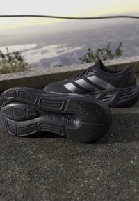 Μαύρα παπούτσια τρεξίματος Adidas με ασημένιες ρίγες, ένα παπούτσι δείχνει την σόλα, τοποθετημένα σε άσφαλτο με θολό μακρινό τοπίο στο φόντο.