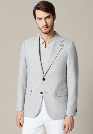 Mężczyzna z brodą ubrany w jasnoszary blezer na szarej koszuli i białe spodnie, stojący na jednolitym beżowym tle.