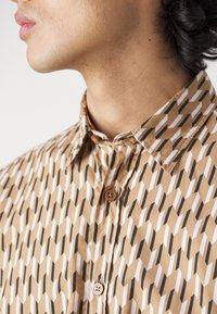 Personne portant une chemise marron, blanche et noire à motifs géométriques, visible du cou jusqu'au milieu de la poitrine.