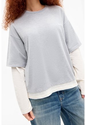Person trägt ein hellgraues übergroßes kurzärmeliges Hemd über einem weißen langärmeligen Shirt und blaue Jeans, steht vor einem weißen Hintergrund.