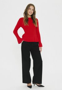 Maglietta rossa a costine con maniche lunghe e polsini svasati, abbinata a pantaloni neri a vita alta e gamba larga e tacchi a punta neri. Sfondo semplice.