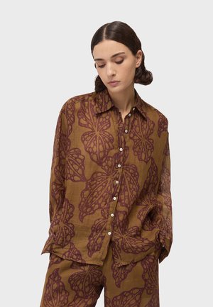 Vrouw die een loszittend bruin shirt en broek draagt met grote donkerrode bladpatronen, naar beneden kijkend met handen in de zakken tegen een effen achtergrond.