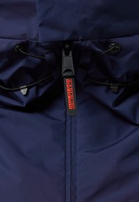 Marineblauwe jas met een hoge kraag, togglesteller en een opvallend rood label. De stof lijkt glad met subtiele textuurdetails.