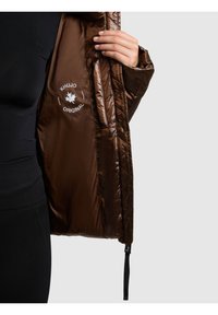 Brązowa pikowana kurtka puchowa z błyszczącą powierzchnią, wewnętrznym nadrukiem logo oraz regulowanym dołem. Dopasowany krój, wysoki kołnierz, miękki materiał i długie rękawy.