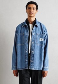 Vaaleansininen farkk shirt, jossa on rento leikkaus, kaksi etutaskua, valkoista tikkausta ja CK-logo-patch vasemmassa taskussa. Napitettu malli.