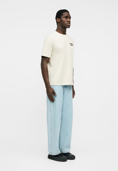 Άνδρας που φοράει κρεμ κοντομάνικη μπλούζα με λογότυπο, φαρδιά τζιν παντελόνια ανοιχτού γαλάζιου χρώματος και μαύρα παπούτσια, στέκεται μπροστά σε απλό λευκό φόντο.