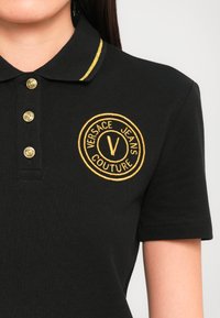 Black polo shirt featuring a gold striped collar, gold buttons, and an embroidered circular logo with "Versace Jeans Couture" text.