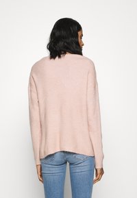 Maglione rosa chiaro a maglia con un fit rilassato e un colletto rotondo. Presenta una texture a coste e un orlo posteriore leggermente più lungo. Indossato con jeans blu.