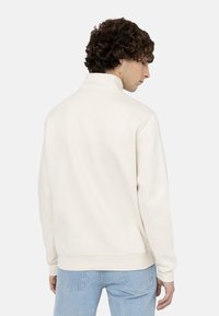 Chaqueta de punto beige claro con un cuello alto, puños y dobladillo acanalados, y una textura suave. Llevada al revés, mostrando el diseño minimalista.
