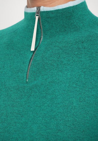Pullover de fleece verde-água com um colarinho alto, apresentando um fecho de correr de metal prateado e um detalhe azul-claro na borda do colarinho. Textura suave.