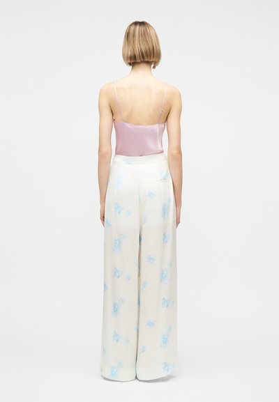 Γυναίκα με κοντά ξανθά μαλλιά, φορώντας ροζ σατέν καμισόλ και ψηλόμεσα, φαρδιά λευκά παντελόνια με μπλε λουλουδάτα σχέδια, στέκεται γυρισμένη προς τα πίσω.