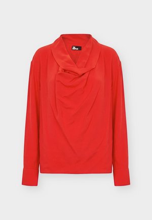 Femme debout face à l’avant, portant un chemisier rouge drapé, un pantalon droit noir et des chaussures noires à bout pointu sur un fond gris clair.