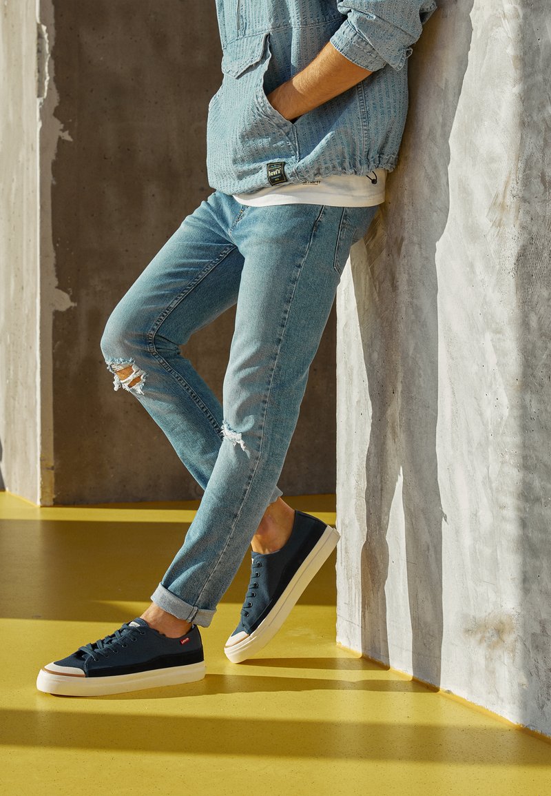 Blaues Jeansjacke aus strukturiertem Stoff, hellblaue, ausgebleichte Jeans und marineblaue Canvas-Sneaker mit weißer Sohle und rotem Logo-Tag.