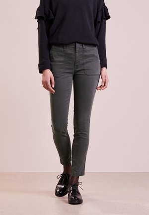 Woman wearing black ruffle-sleeve top, fitted dark gray pants with front pockets, black sheer tights, and black shiny lace-up shoes.