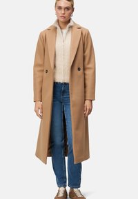 Long manteau en laine couleur camel avec des revers et des boutons doubles, superposé sur un pull texturé crème et un jean bleu. Chaussures décontractées marron.