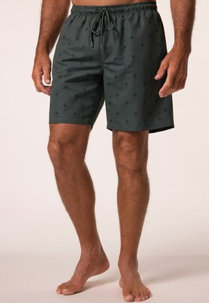 Mann trägt dunkelgrüne Badeshorts mit kleinen schwarzen Palmenmustern und Kordelzug am Bund, steht barfuß vor einem schlichten Hintergrund.