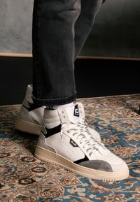 Białe skórzane sneakersy typu high-top z szarymi zamszowymi akcentami, czarnymi wstawkami i wzorzystymi sznurówkami. Posiadają logo na języku i perforacje.