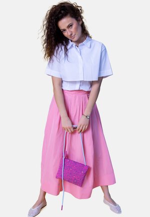 Woman with curly hair wearing a white cropped shirt, pink midi skirt, holding a pink leopard print purse with long blue straps.
