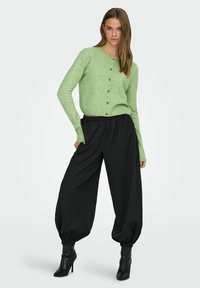 Roheline kootud kardigan, millel on nööbid, kombineeritud lahtiste mustade pükstega ja mustade kontsakingadega. Sile tekstuur, lõdvestunud lõige.