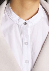 Vit skjort med krage som har rundad nacke, silverfärgad tryckknapp och strukturväv. Bakgrunden visar ett mjukt beige material.