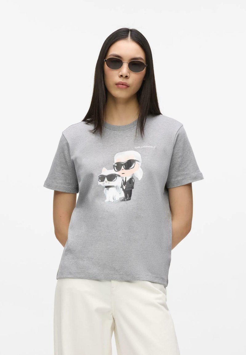 Femme aux longs cheveux foncés portant de petites lunettes de soleil ovales, un t-shirt gris avec des personnages dessinés de Karl Lagerfeld et un chien, et un pantalon blanc.