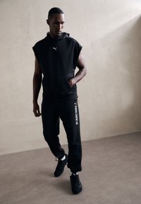 Zwart mouwloos hoodie en sweatpants outfit, met een zak aan de voorkant en witte logo-details, gedragen met zwarte sportschoenen. Vlakke textuur.