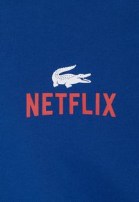 Modré bavlněné tričko s bílou grafikou krokodýla a slovem "NETFLIX" tučně červeně napsaným těsně pod ním. Hladká textura.