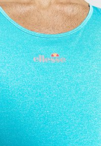 Uomo che indossa una canottiera sportiva azzurro chiaro con il logo ellesse al centro del petto, mostrando parte del collo peloso e della parte superiore del petto.