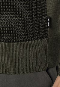 Tummanvihreä neulepaita, jossa teksturoitu ja kuvioitu yläosa sekä resoroitu helma. Pienessä mustassa kylkietiketissä lukee "BOSS".