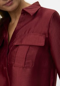 Maroon shirt in a lightweight, textured fabric features a collar, two front pockets, and a buttoned placket. Elegant stripe pattern visible.