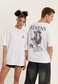 Jonge vrouw en man dragen witte T-shirts met grafische afbeeldingen van Athene; op de voorkant van het shirt van de vrouw staat een klein logo, op de achterkant van het shirt van de man een groot ontwerp met Grieks thema.