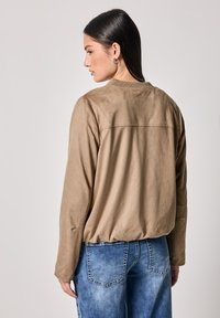 Giacca in suede tan con maniche lunghe e colletto a costine, abbinata a jeans blu sfumati. Texture morbida e vestibilità rilassata, che mette in evidenza un design semplice.