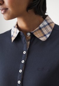 Femme portant une chemise bleu marine avec des boutons blancs et un col à carreaux beige, bleu et blanc, montrant la partie supérieure de la poitrine et du cou.