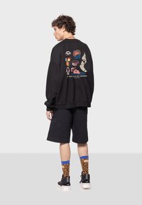 Sudadera negra con impresiones gráficas coloridas en la parte posterior, combinada con pantalones cortos negros y calcetines brillantes con estampado de leopardo. Zapatillas negras completan el look.