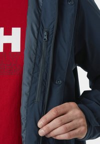 Giacca blu navy con tessuto liscio, tasca con zip e dettagli in bottone. Camicia rossa sotto con grandi lettere bianche.