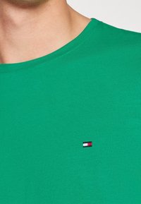 Zielona bawełniana koszulka z okrągłym dekoltem i krótkimi rękawami, z małym wyszywanym logo w kolorze czerwonym, białym i niebieskim na piersi.