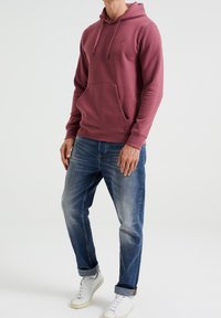 Mörkröd huvtröja med ficka framtill, kombinerad med urtvättade blå jeans och vita sneakers. Slät textur, justerbara dragsnören på huvan.