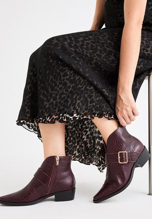 Botines burdeos con un patrón de cocodrilo texturizado, punta afilada, cremallera lateral y detalle de hebilla decorativa, combinados con una falda de estampado de leopardo.