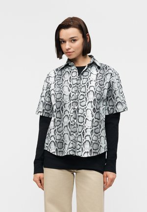 Jauna moteris su tiesiais rudais plaukais, dėvinti trumpomis rankovėmis pilką marškinėlį su gyvatės raštu, dėvimą ant juodos ilgarankovės palaidinės ir smėlio spalvos kelnes.