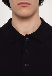 Lähelle otettu kuva henkilön alahuulesta ja kaulasta, jolla on päällään teksturoitu musta nappipaita, jossa on leveä kaulus.