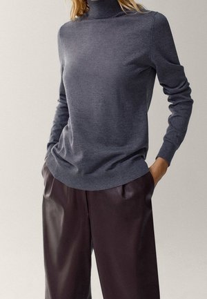 Personne portant un pull gris à col roulé ajusté et un pantalon en cuir marron foncé ample, les mains dans les poches.