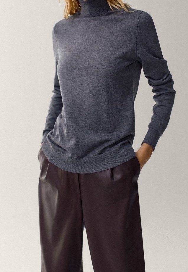 Personne portant un pull gris à col roulé ajusté et un pantalon en cuir marron foncé ample, les mains dans les poches.