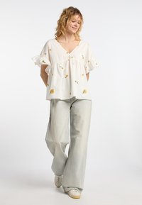 Blusa di cotone bianca con ricami floreali gialli, scollatura a V e maniche corte con volant, abbinata a jeans a gamba larga blu chiaro e sneakers bianche.