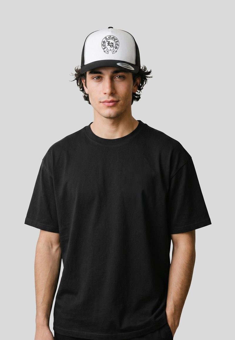 Jeune homme aux cheveux bouclés foncés porte un t-shirt noir et une casquette de baseball blanche et noire avec un motif circulaire en forme de fer à cheval.