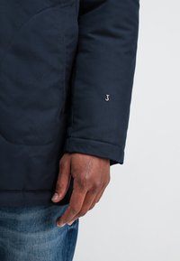 Donkerblauwe gewatteerde jas met een gladde textuur, voorzien van een zilveren "J"-logo op de mouw en een standaard manchetontwerp.