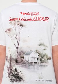 Λευκό βαμβακερό μπλουζάκι με γραφικό που απεικονίζει ένα σπίτι δίπλα στη λίμνη και δέντρα, με την επιγραφή «Serene Lakeside LODGE» σε κόκκινο και λεπτομερή σχεδίαση.