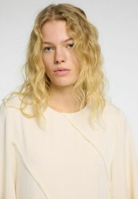 Blusa de color crema con mangas largas, presenta una costura diagonal con bordes deshilachados, made de una tela liviana y texturizada.