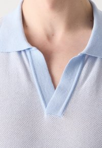 Vue en gros plan du cou et de la poitrine d'une personne portant un polo bleu clair texturé à col avec un décolleté en forme de V.