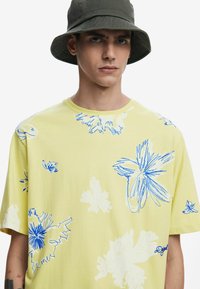 Camiseta amarilla de manga corta con patrones florales en azul y blanco. Tejido suave con cuello redondo y corte relajado.