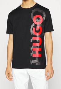 Sort bomulds T-shirt med et fremtrædende, stiliseret "HUGO" logo i rød og grå, med rund halsudskæring og korte ærmer.
